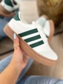 Tenis Addi blancos con detalles verdes, suela color café y tres líneas laterales verdes. Diseño cómodo, versátil y moderno para uso diario.